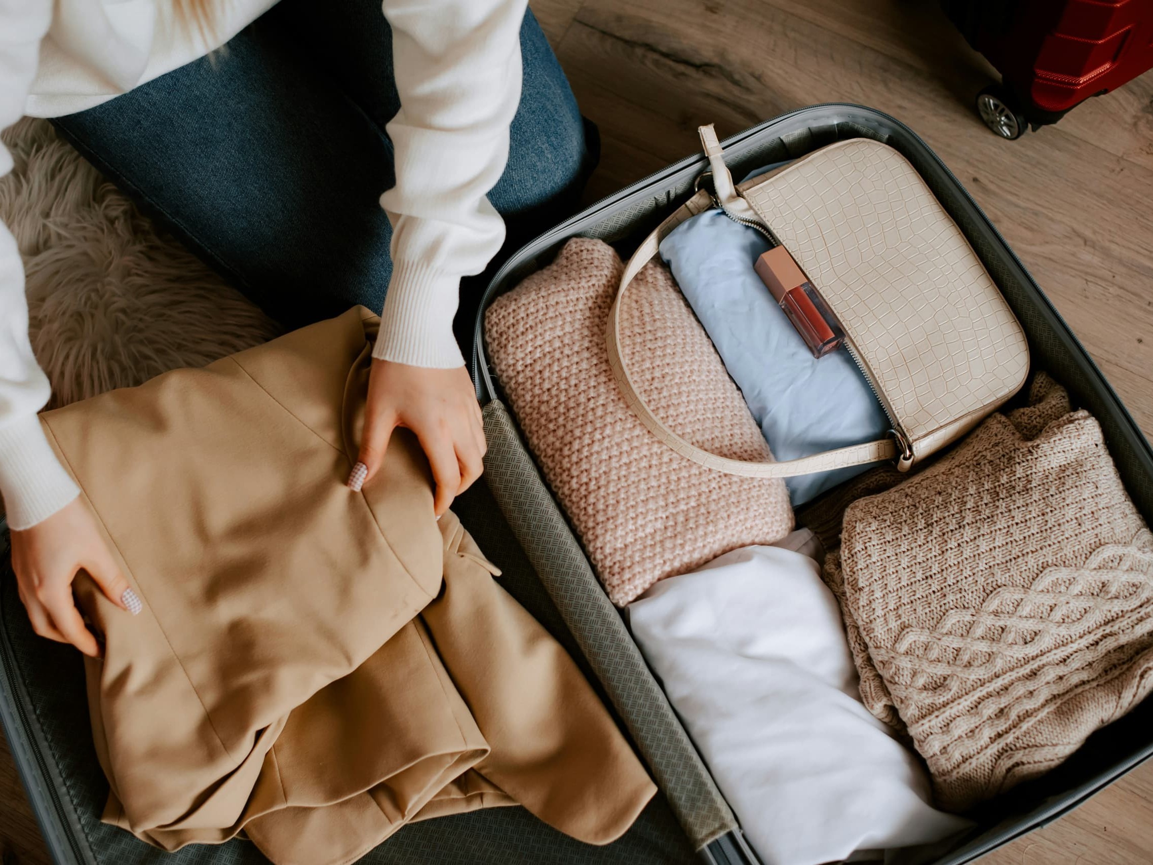 Person kneeling beside an open suitcase, packing a beige coat, knitted blankets, and a light blue garment inside.
