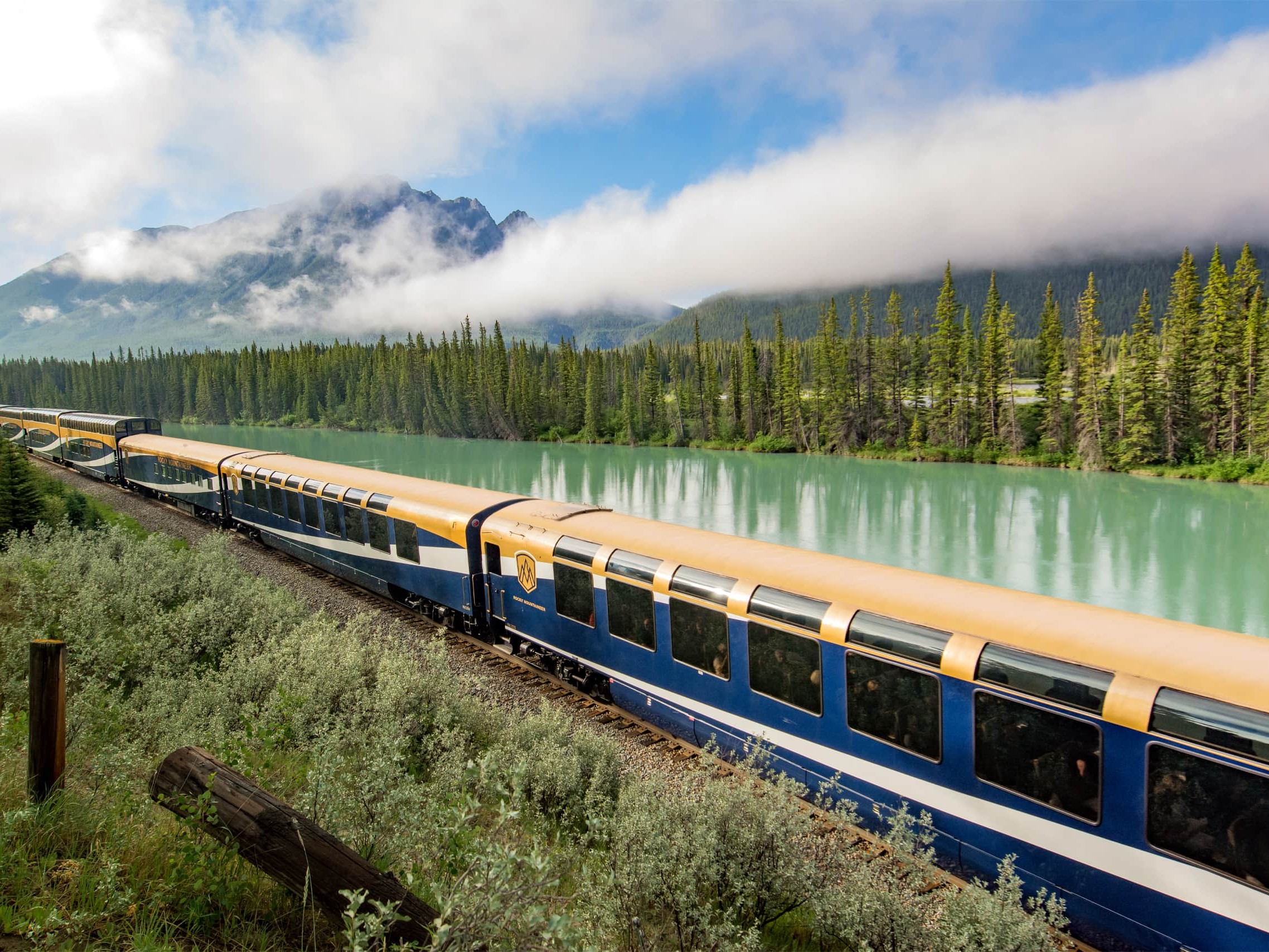 Blue-and-orange passenger train winds along a turquoise river with evergreen trees and misty mountains in the background.