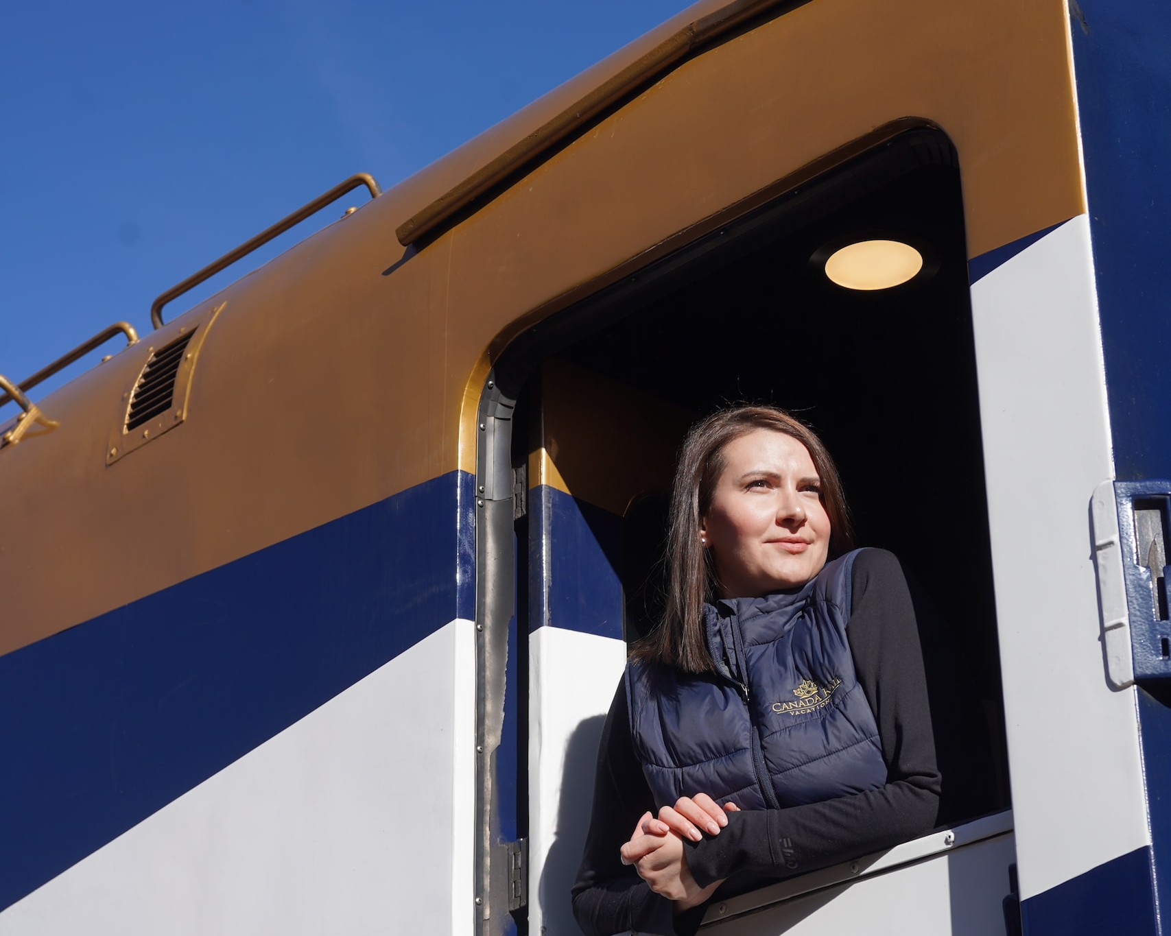 Woman leaning out from a train cab door on a blue and tan locomotive, against a clear blue sky.