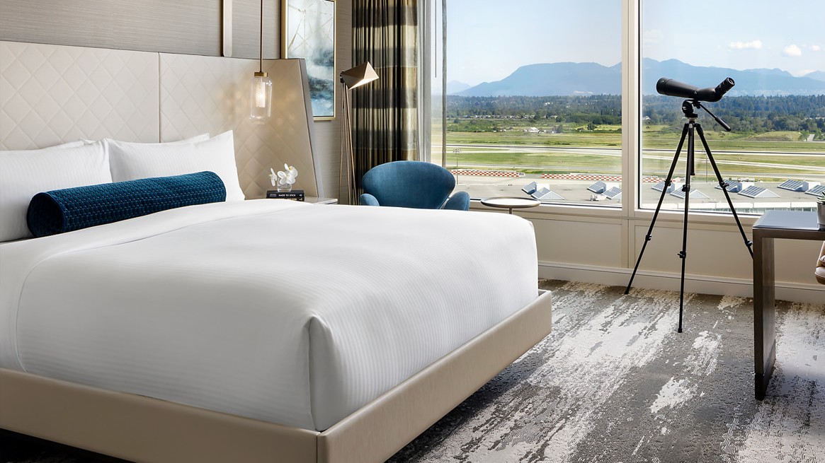 Hotel room with a large bed and blue bolster, beside a tripod telescope by a window overlooking mountains and a runway.