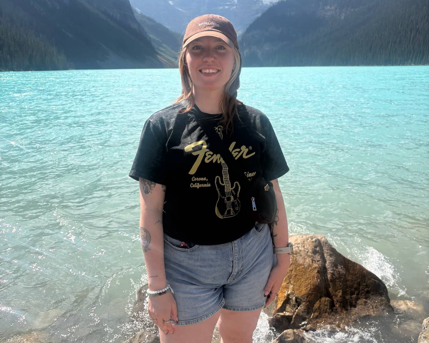 Smiling woman standing on rocks at a turquoise alpine lake with snow-capped mountains in the background.