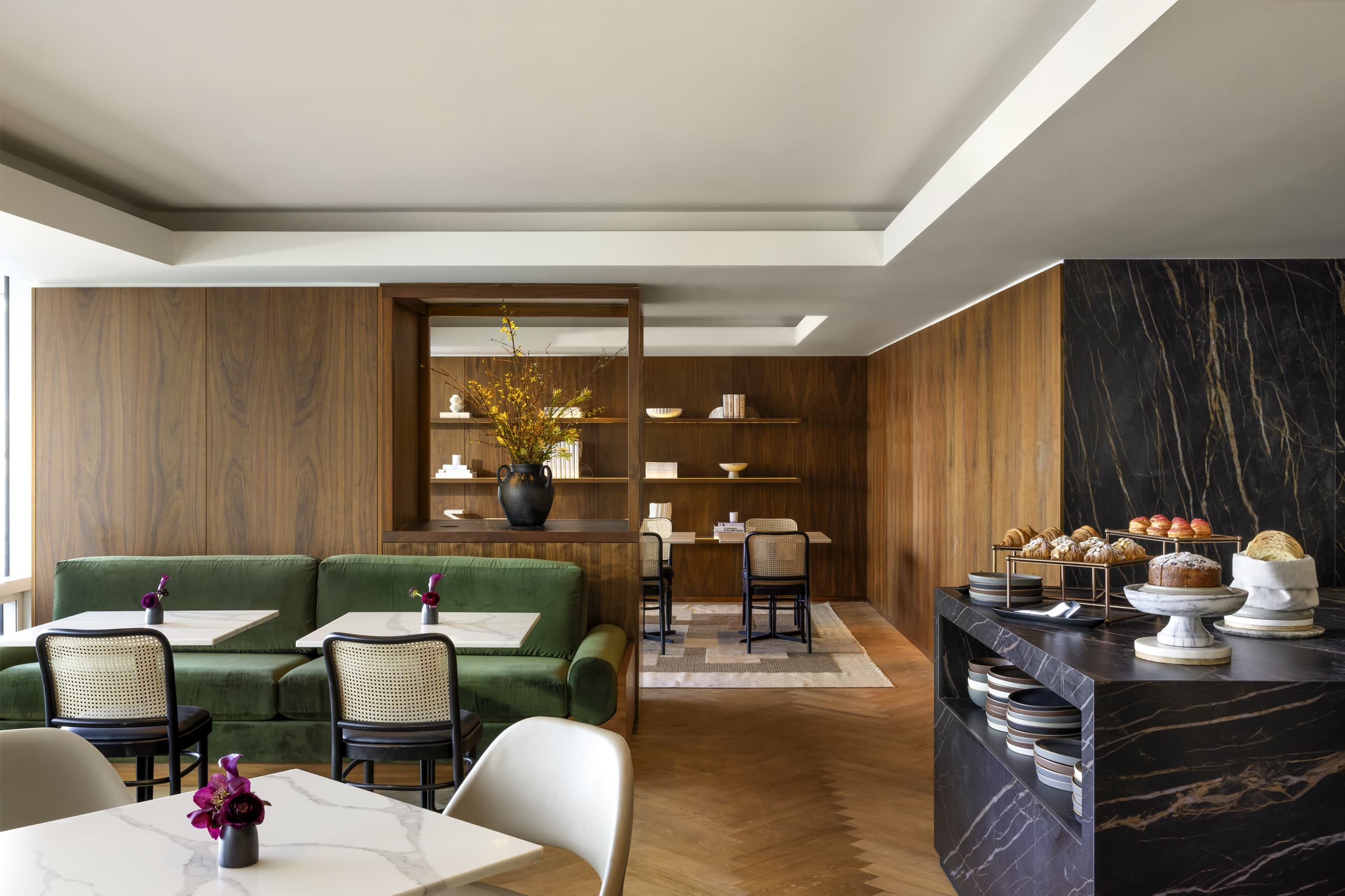 Cafe interior with warm wood paneling, green sofa seating, and marble-topped tables in foreground; pastries displayed on a black marble counter to the right.