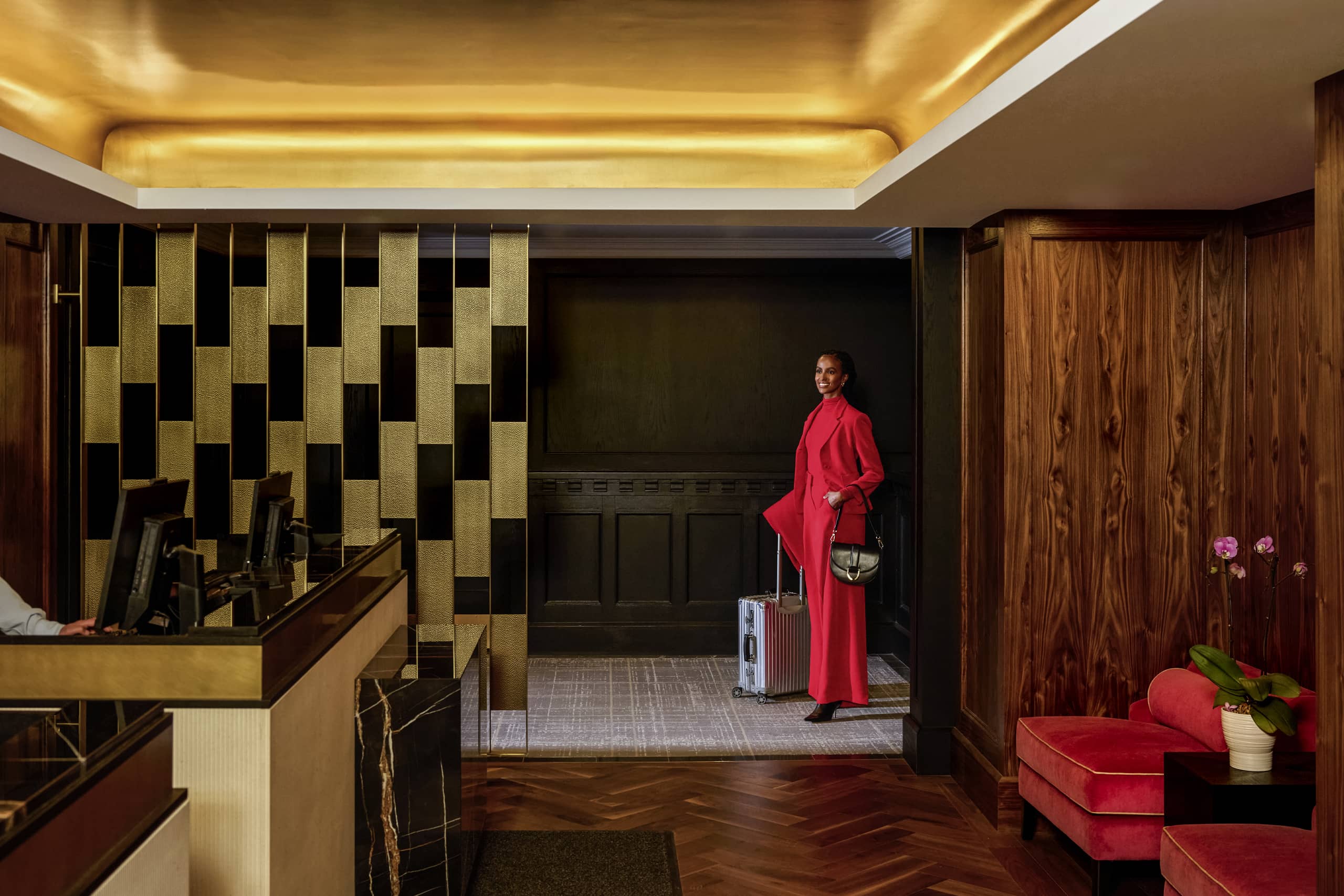 Smiling woman in a red coat standing with a suitcase in a gold-accented hotel lobby near the reception desk.