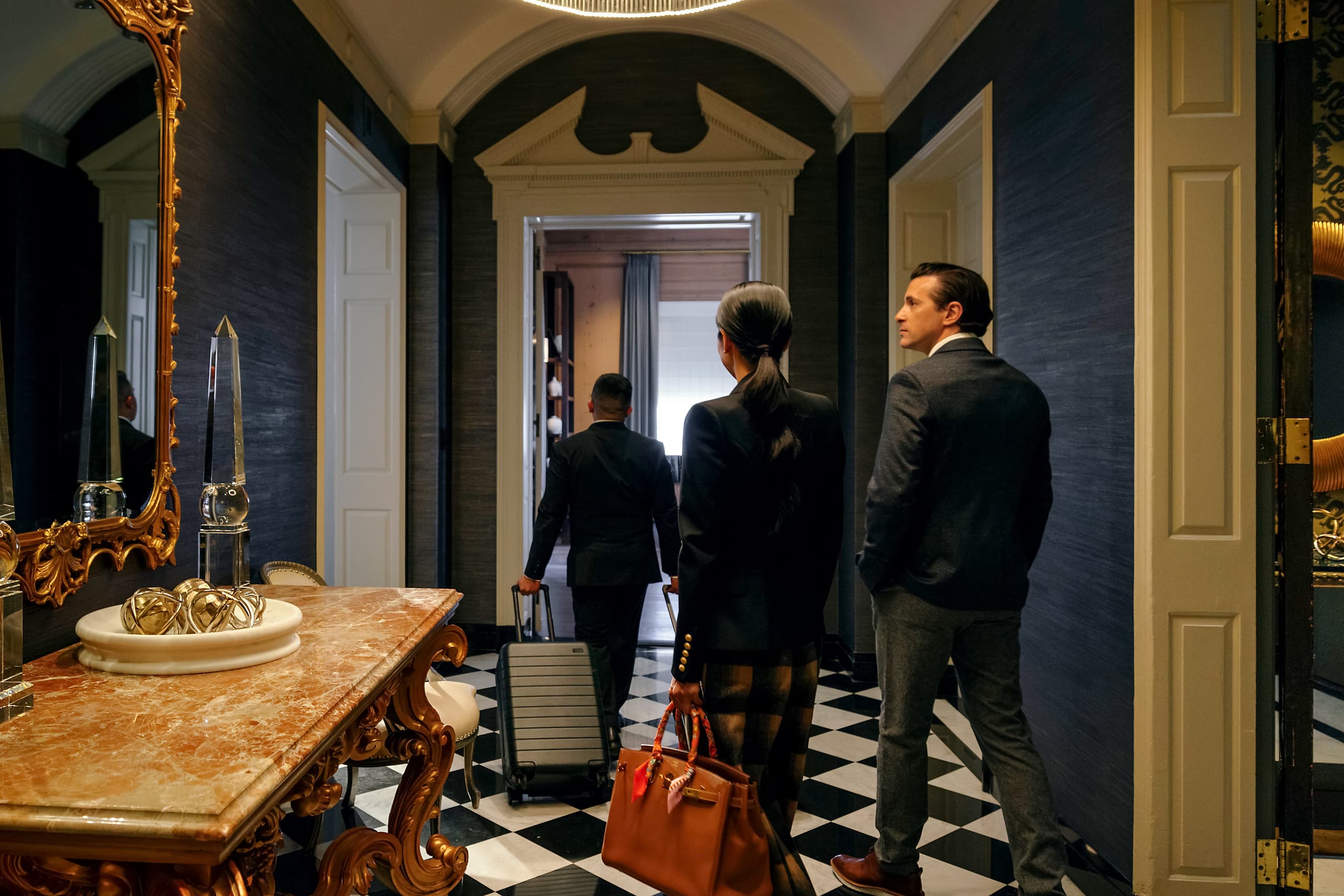 Three professionals in suits with rolling luggage stand in a luxurious hallway with a checkered floor and a gold-framed mirror nearby.