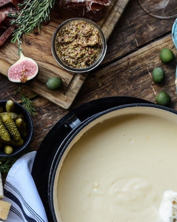 Creamy cheese fondue pot with bread cubes, pickles, olives, and mustard on a rustic wooden table with herbs