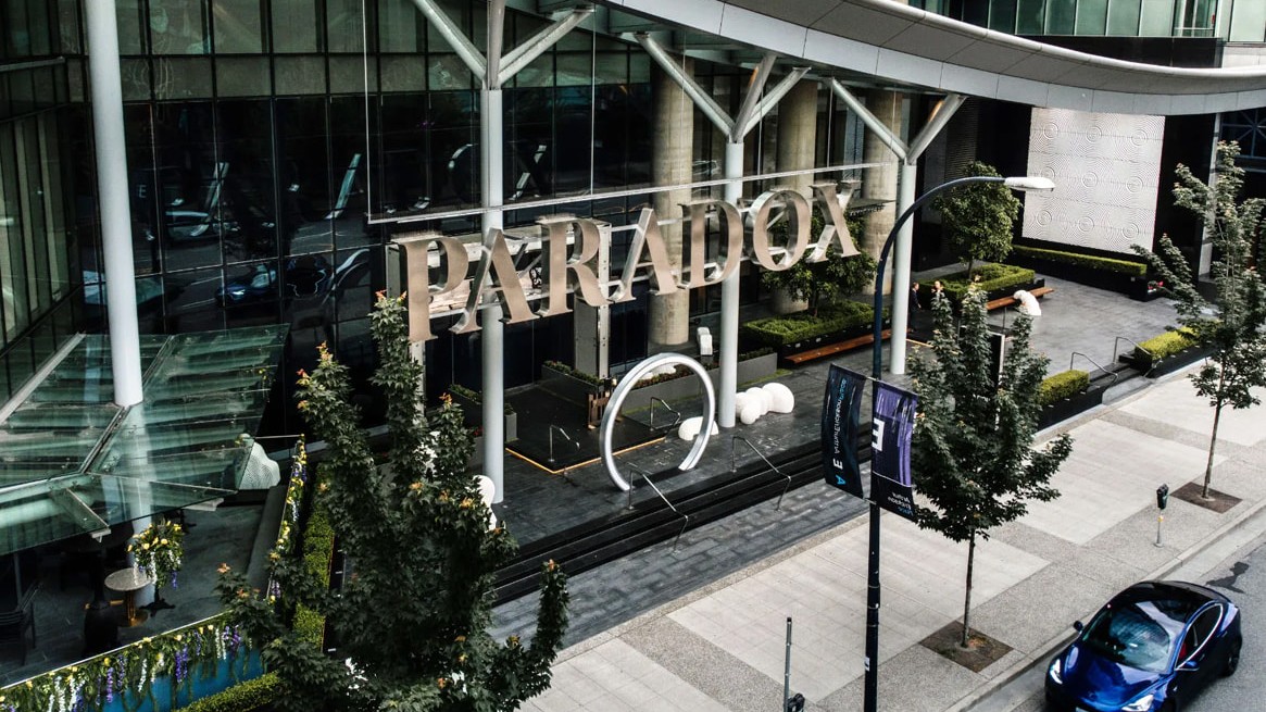 Aerial view of a glass-front building with a large PARADOX sign over the entrance, trees and a blue sports car on the street.