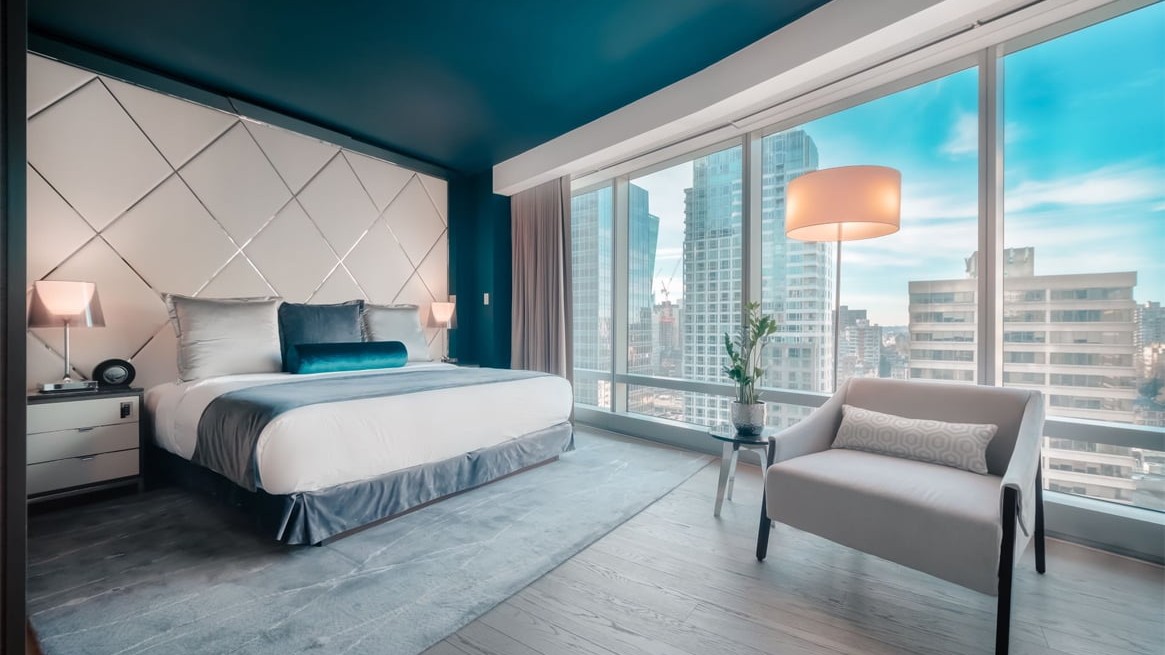Modern hotel bedroom with a king bed, blue-gray decor, and skyline view through floor-to-ceiling windows.